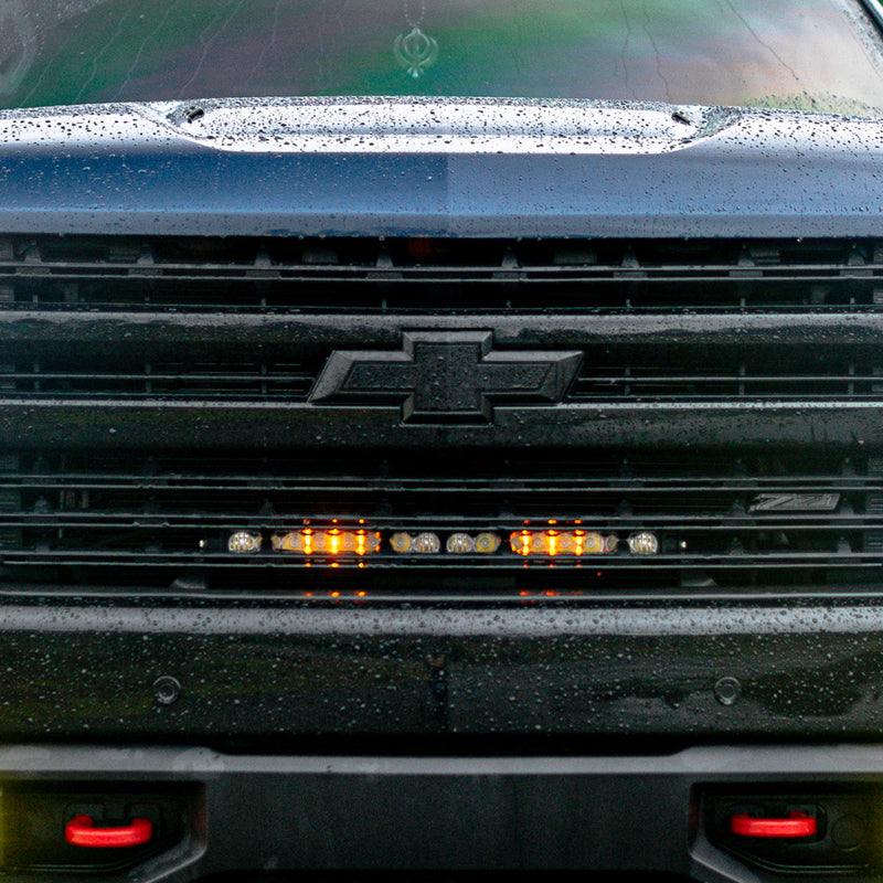 Baja Designs S8 20" Grille Light Bar Kit on a Chevrolet Silverado 1500: front view of a black Chevrolet Silverado grille with a lit light bar glowing behind the slats below the bowtie
