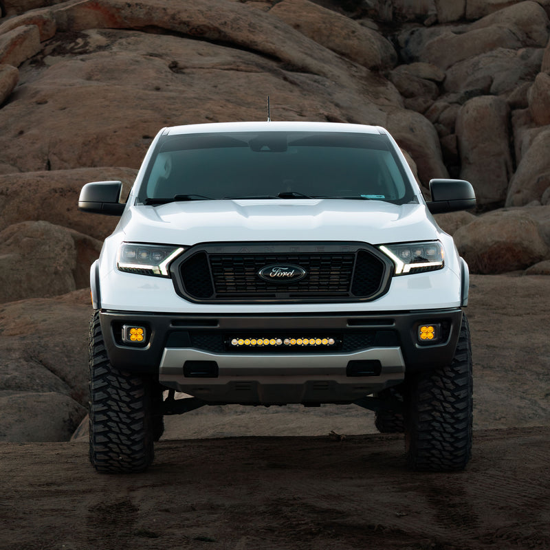 Baja Designs S8 20" Grille Light Kit installed on a Ford Ranger: A white Ford Ranger parked among boulders with an amber-lit light bar in the lower grille and amber pods in the fog pockets