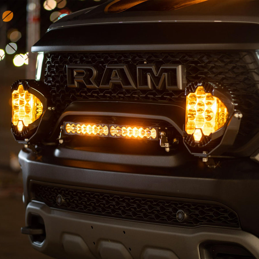 Baja Designs S8 20" Grille Light Kit on a Ram 1500 TRX: Ram TRX grille at night with a short LED light bar and two pods glowing amber below the RAM lettering