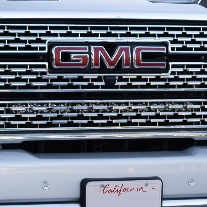 Baja Designs S8 30" Behind the Grille Light Bar Kit on a GMC Sierra HD: close view of a chrome GMC Sierra HD grille with an LED light bar visible behind the lower mesh and a blank California plate below
