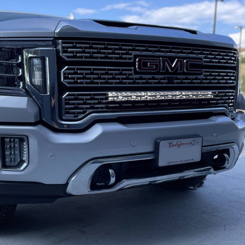 Baja Designs S8 30" Behind the Grille Light Bar Kit on a GMC Sierra HD: silver GMC Sierra HD with a lit LED light bar set behind the lower grille and a blank California plate on the bumper