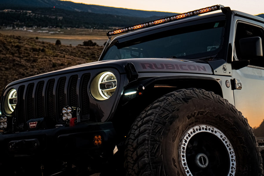Baja Designs S8 50" Roof Mount Light Kit on a Jeep Wrangler JL: A black Jeep Wrangler Rubicon at dusk shows a slim LED light bar glowing above the windshield and pods on the bumper