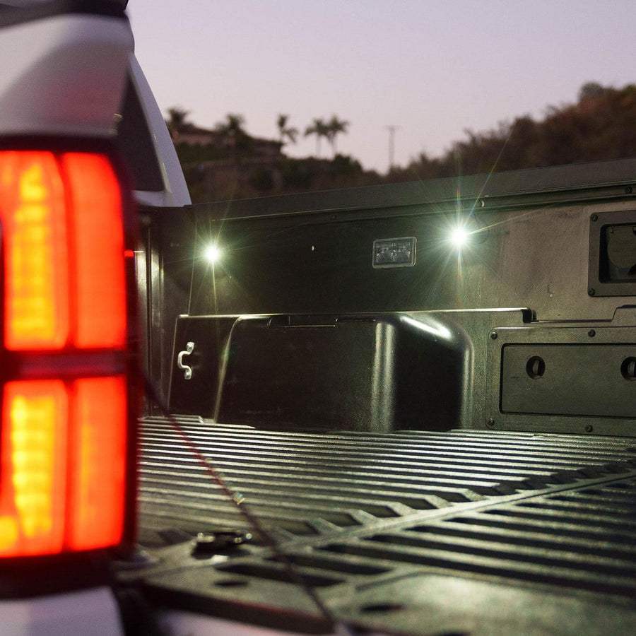 Vehicle Light Mount Kits by Baja Designs: A pickup truck bed lit white by rail-mounted cargo lights at dusk beside a glowing red tail light.