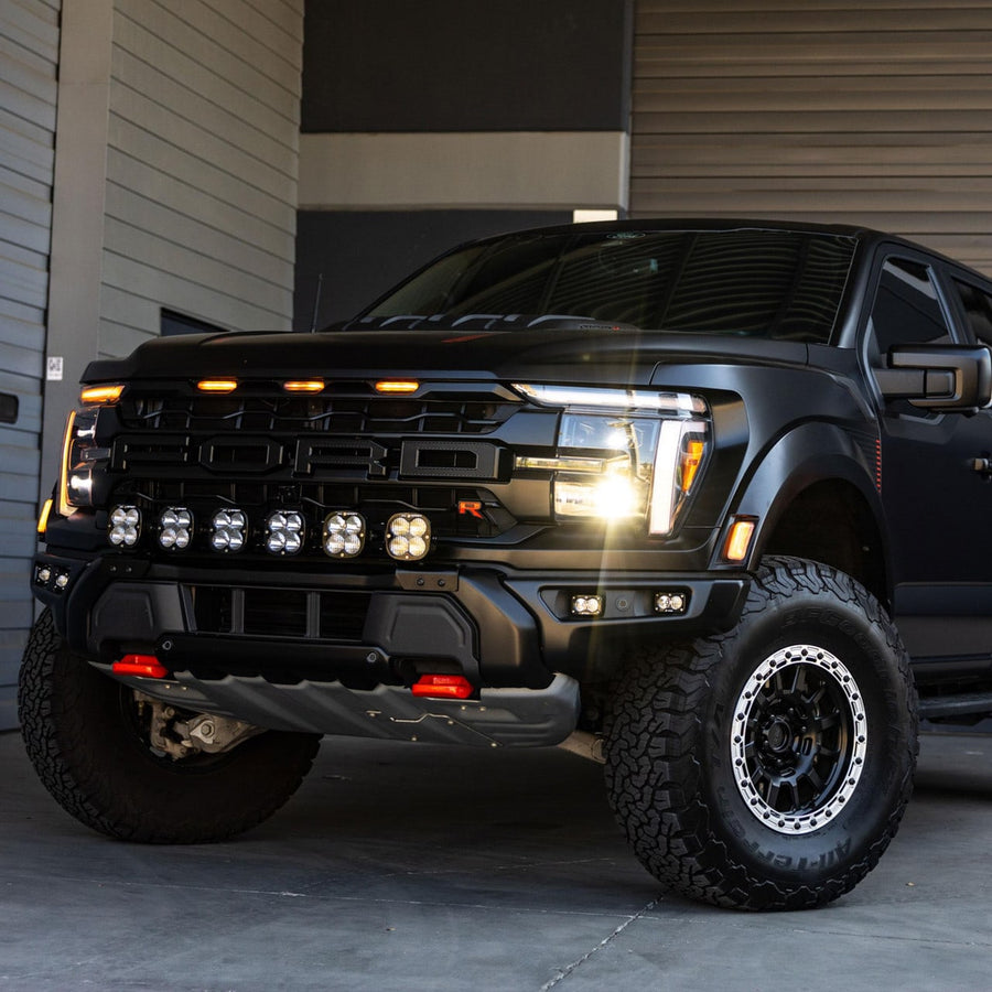 Bumper Light Kits by Baja Designs: A black Ford Raptor pickup parked by a garage door with six linkable LED pods mounted low in the front bumper opening.