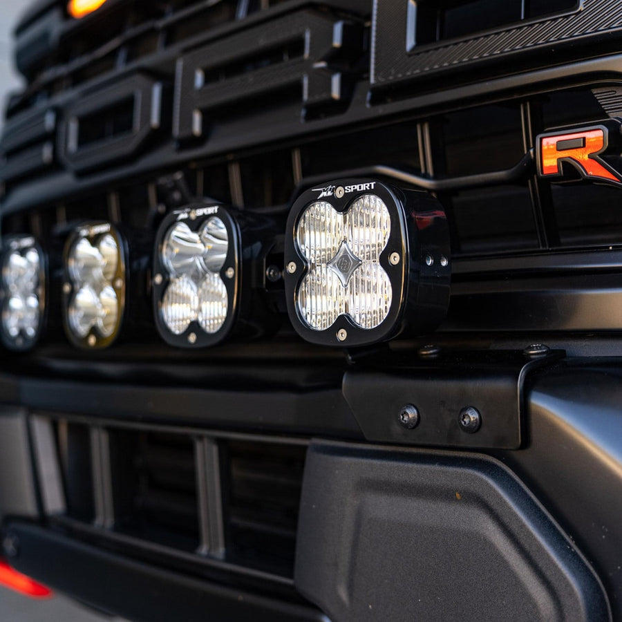 Bumper Light Kits by Baja Designs: Angled close-up of XL Sport linkable pods mounted in the front bumper opening of a black Raptor beside the red R badge.