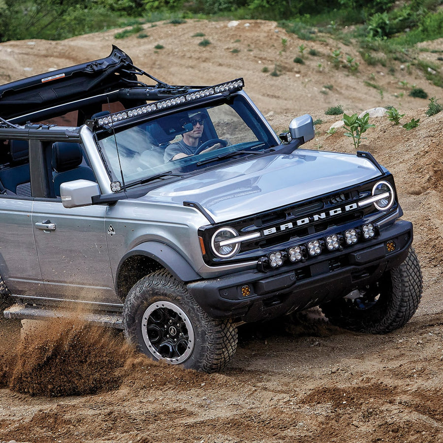 Bumper Light Kits by Baja Designs: A silver Ford Bronco climbs a dirt trail with a six-pod linkable light bar mounted across its front bumper.