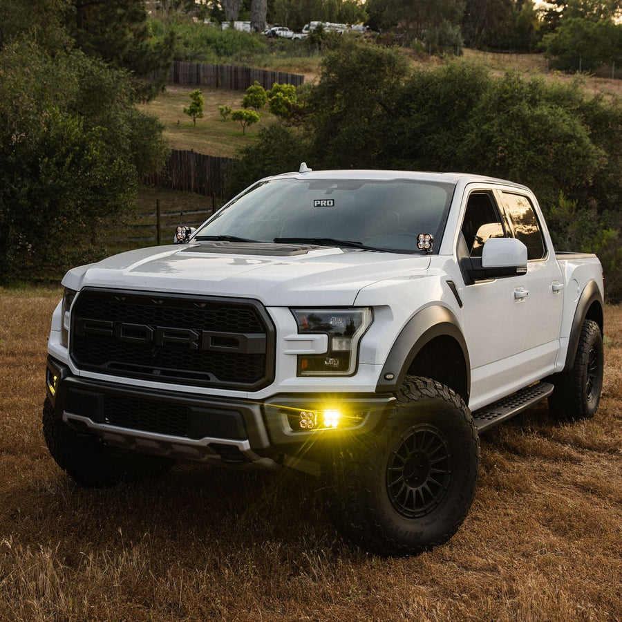 Fog Light Kits by Baja Designs: White Ford Raptor pickup parked on dry grass with amber fog pocket lights lit.