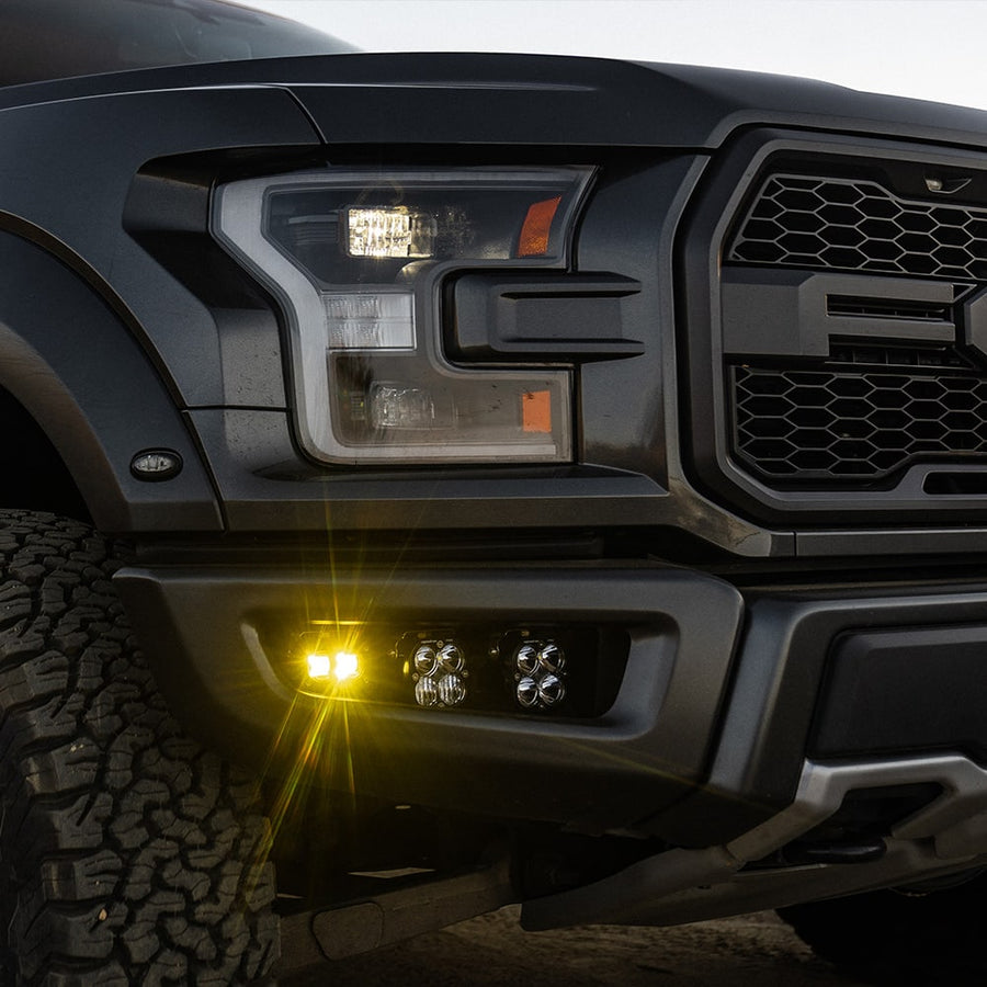 Fog Light Kits by Baja Designs: Close view of a black Ford Raptor front with an amber S2 pod and two clear Squadron pods lit in the fog pocket.