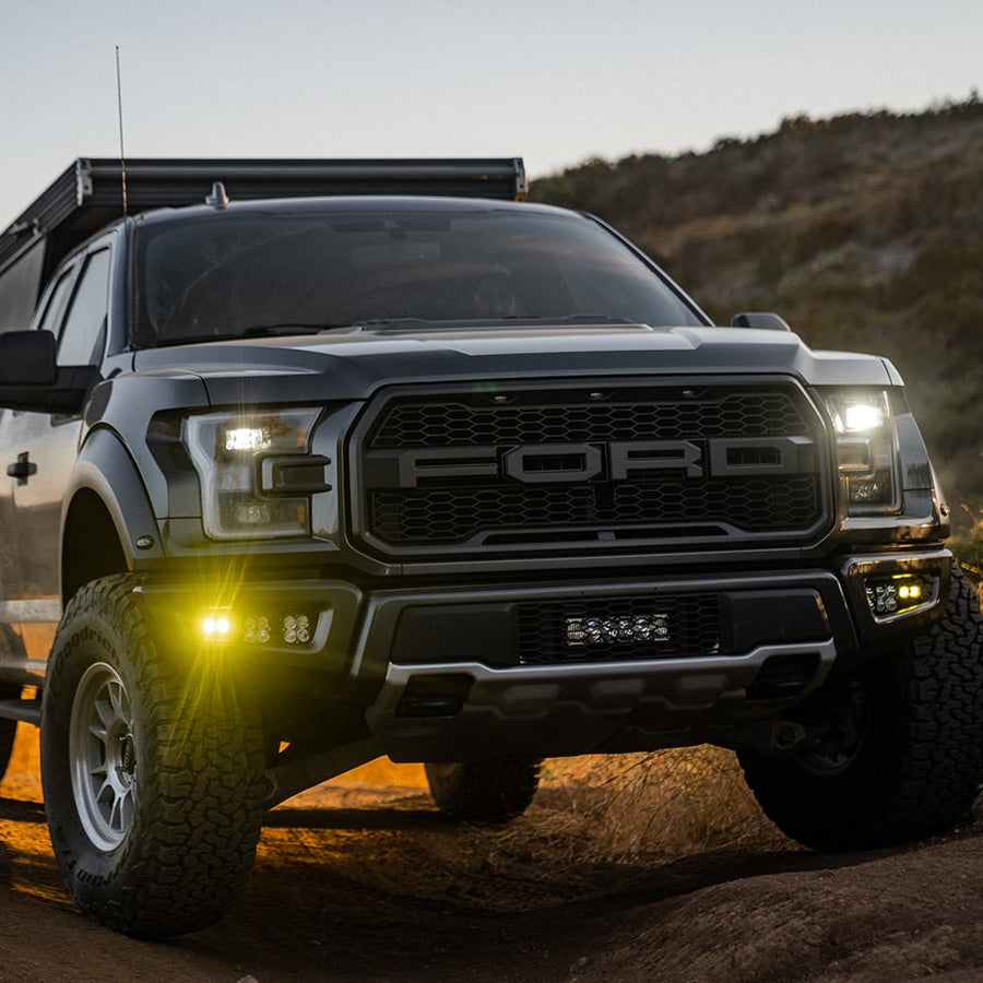 Fog Light Kits by Baja Designs: A black Ford Raptor on a dirt trail at dusk with amber fog pocket lights glowing in the bumper.