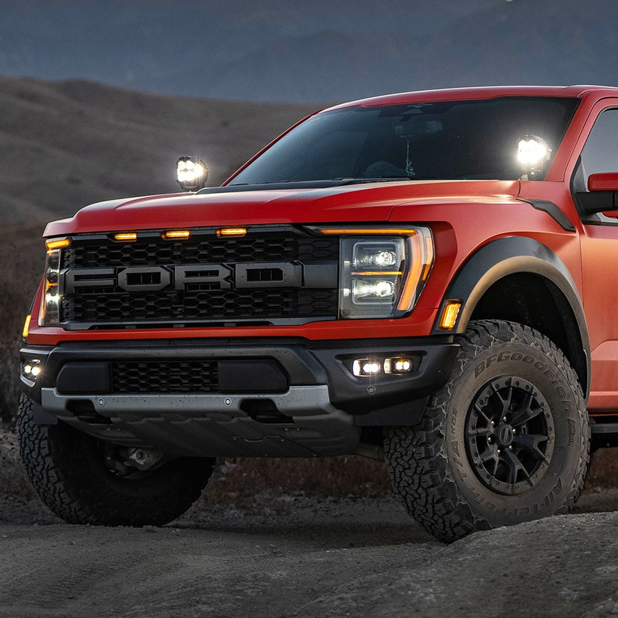 Fog Light Kits by Baja Designs: A red Ford Raptor at dusk with lit fog pocket lights in the bumper and extra pods lit near the windshield.