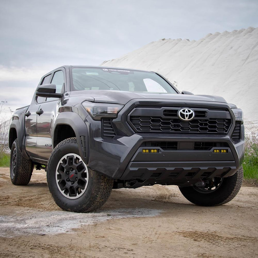 Fog Light Kits by Baja Designs: A gray Toyota Tacoma pickup on a dirt lot with amber S2 pods fitted in its lower bumper fog pockets.