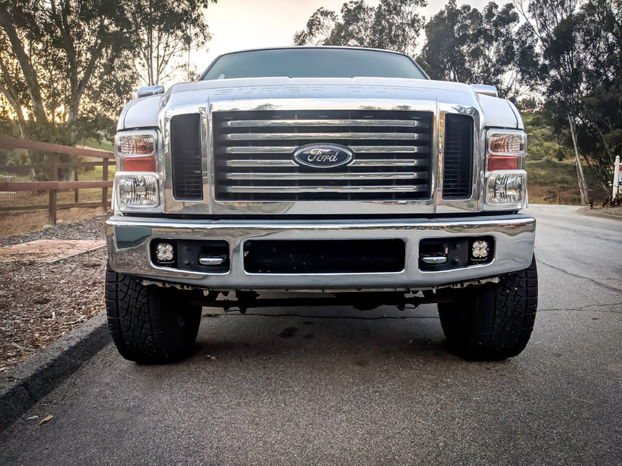 Fog Light Kits by Baja Designs: Front view of a silver Ford Super Duty pickup on a paved road with Squadron pods in its bumper fog pockets.