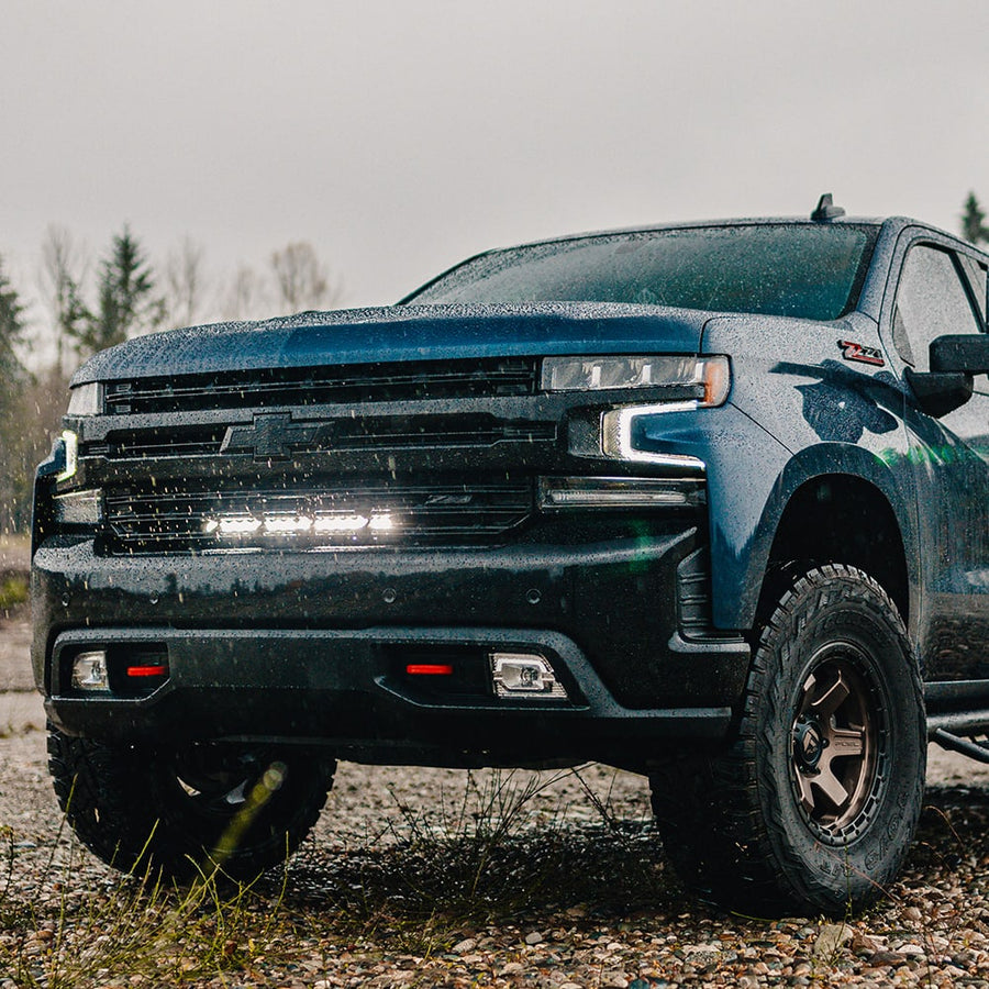 Grille Light Kits by Baja Designs: A dark blue Chevrolet Silverado parked on a gravel trail with a lit light bar glowing in the grille.