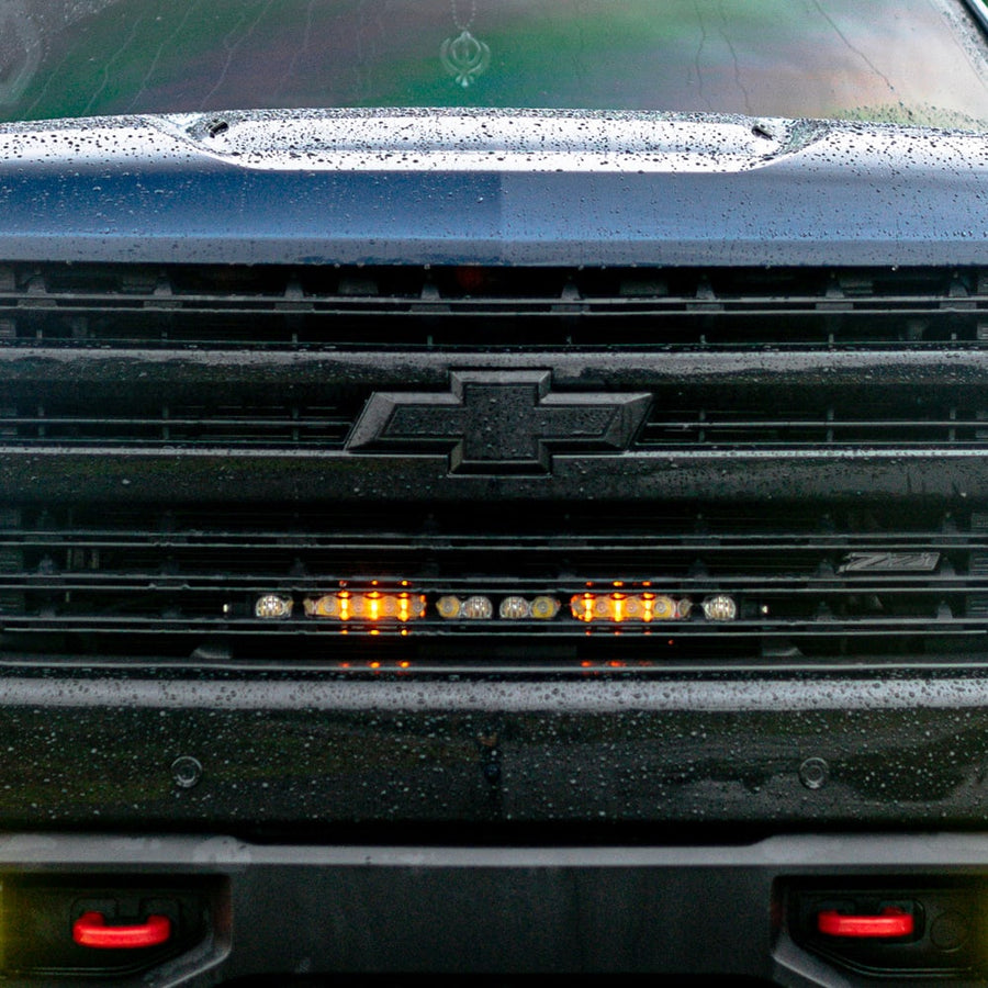 Grille Light Kits by Baja Designs: Front view of a black Chevrolet Silverado grille with a lit light bar glowing behind the slats below the bowtie.