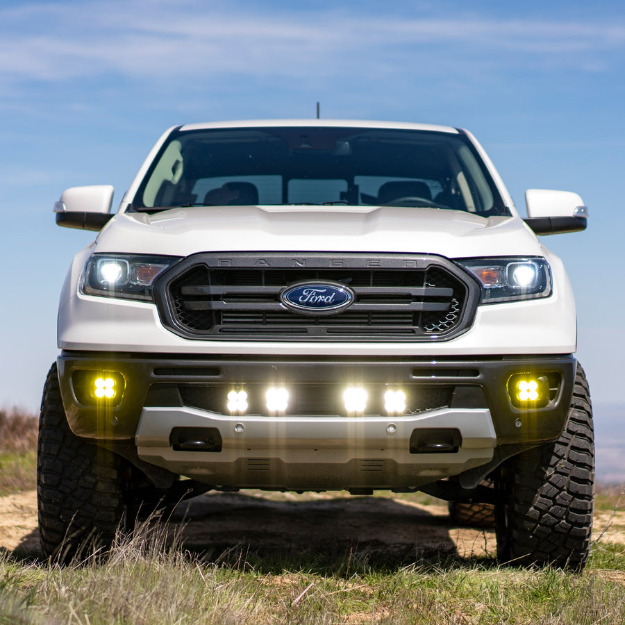 Grille Light Kits by Baja Designs: A white Ford Ranger under blue sky with four lit square pods in the lower grille and amber pods in the fog pockets.