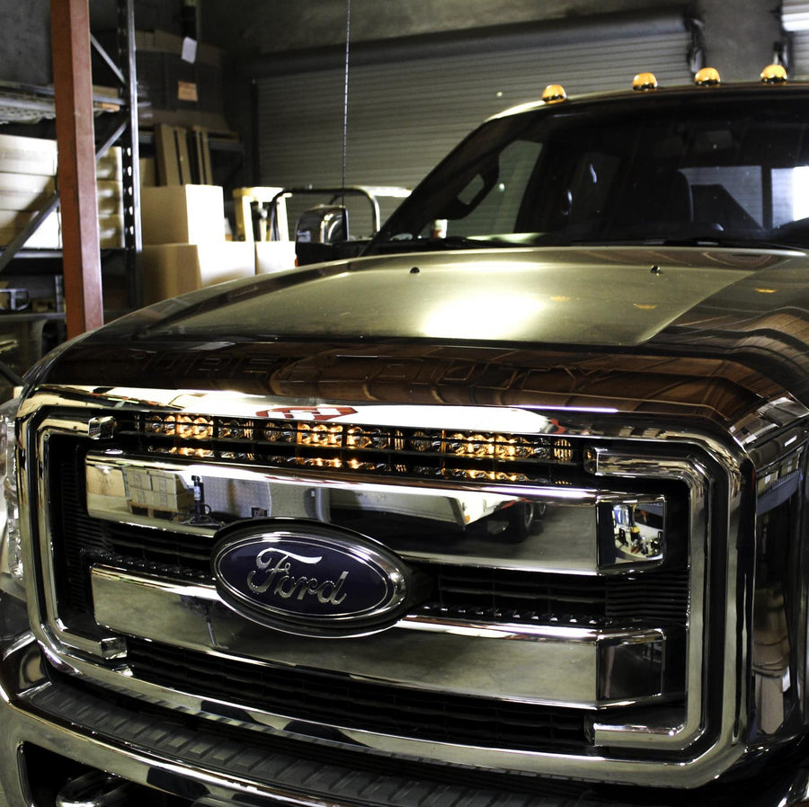 Grille Light Kits by Baja Designs: Dark Ford Super Duty in a shop with a lit LED light bar mounted across the upper grille above the Ford oval.