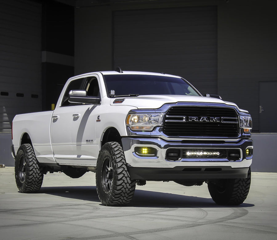 Grille Light Kits by Baja Designs: White Ram 2500 in front of a warehouse with a lit LED light bar in the lower bumper grille and amber fog lights.
