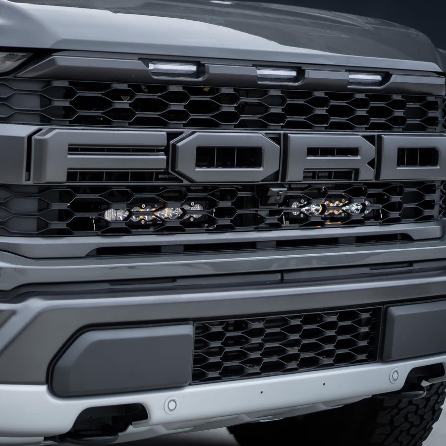 Grille Light Kits by Baja Designs: Gray Ford Raptor grille with two short LED light bars mounted behind the lower grille below the FORD lettering.