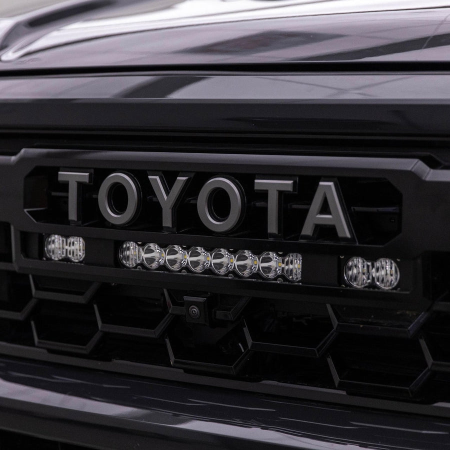 Grille Light Kits by Baja Designs: A clear lens 10 inch S8 bar between two S2 Pro pods mounted under the TOYOTA lettering of a dark Tacoma grille.