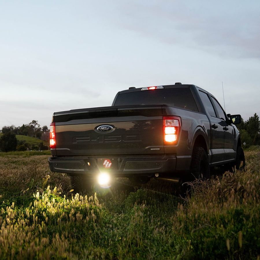 Rear & Utility Lights by Baja Designs: A dark Ford F-150 seen from the rear on a grassy field at dusk with a hitch light glowing below the tailgate.