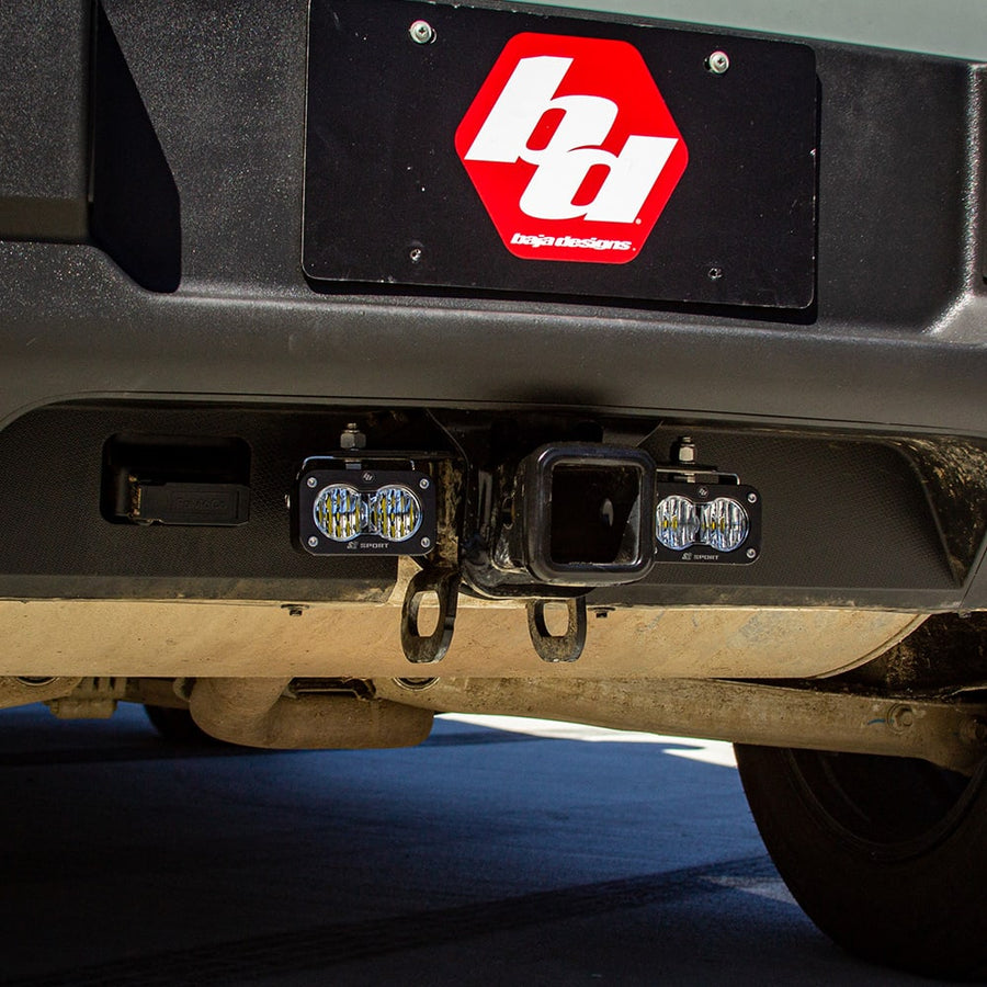 Reverse Light Kits by Baja Designs: Two S2 pods mounted on either side of a receiver hitch below a rear bumper with a Baja Designs logo plate.