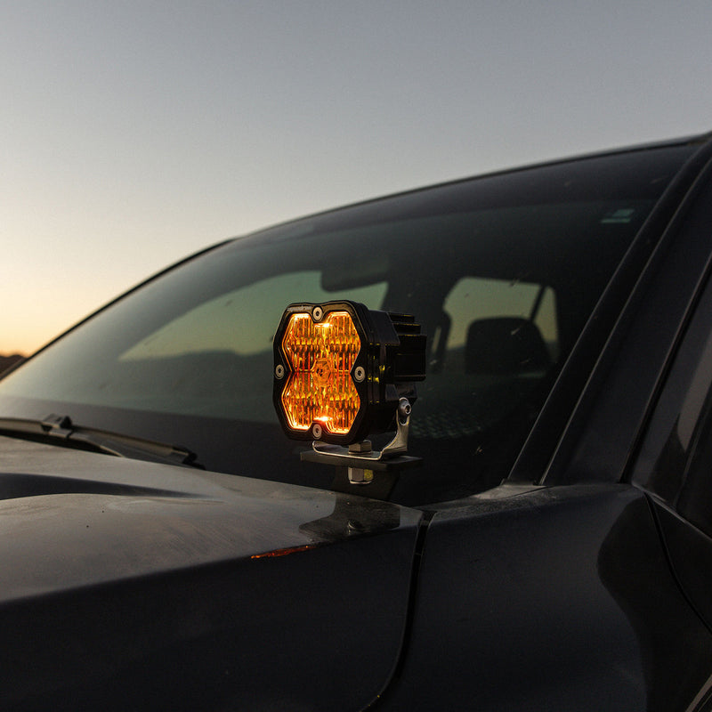 Baja Designs Squadron 2.0 Pro A-Pillar Light Kit on a vehicle: A Squadron pod lit amber on an A-pillar bracket at the base of a dark vehicle windshield
