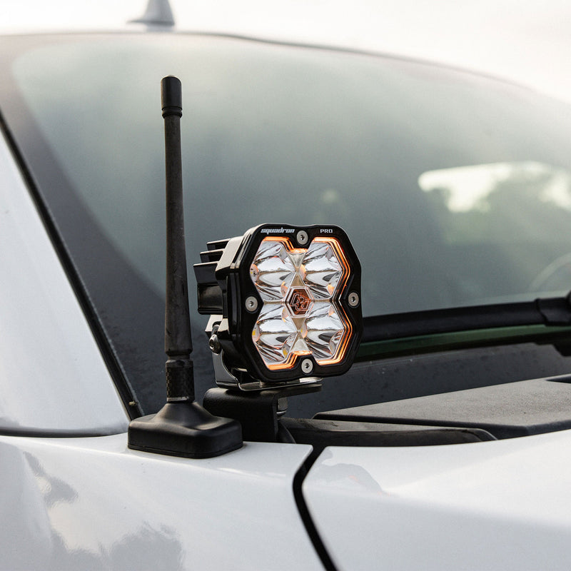 Baja Designs Squadron 2.0 Pro A-Pillar Light Kit on a vehicle: A Squadron pod with a glowing amber backlight on an A-pillar bracket beside a short antenna on a silver vehicle