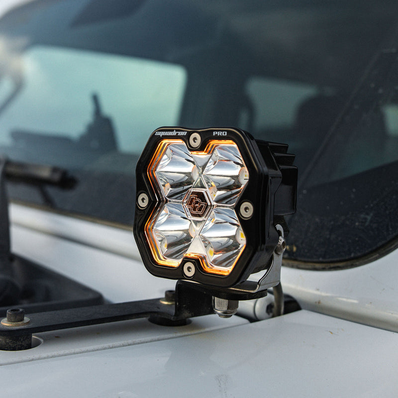 Baja Designs Squadron 2.0 Pro A-Pillar Light Kit on a Jeep Wrangler: A Squadron Pro pod with an amber backlight on a cowl mount beside the windshield of a white Jeep