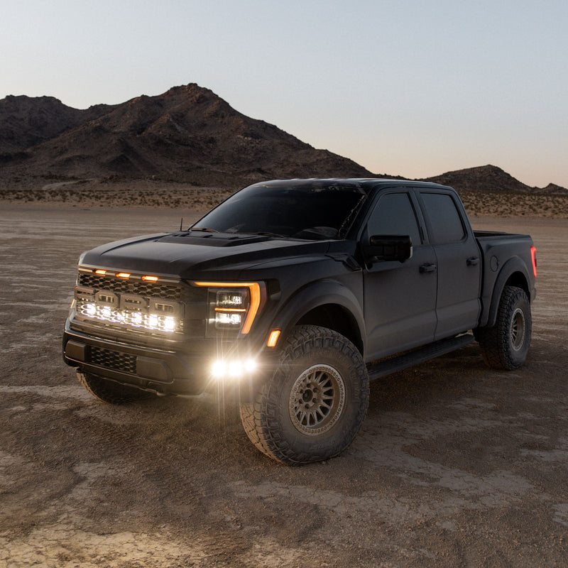 Baja Designs Squadron 2.0 Pro Behind-the-Grille Light Kit installed on a Ford F-150 Raptor: A black Ford Raptor on a desert flat at dusk with lit pods behind its grille and in the bumper