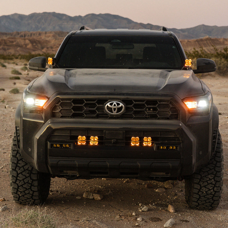 Baja Designs Squadron 2.0 Pro Behind-the-Grille Light Kit installed on a Toyota: front view of a dark Toyota in the desert at dusk with four lit amber pods behind the lower grille