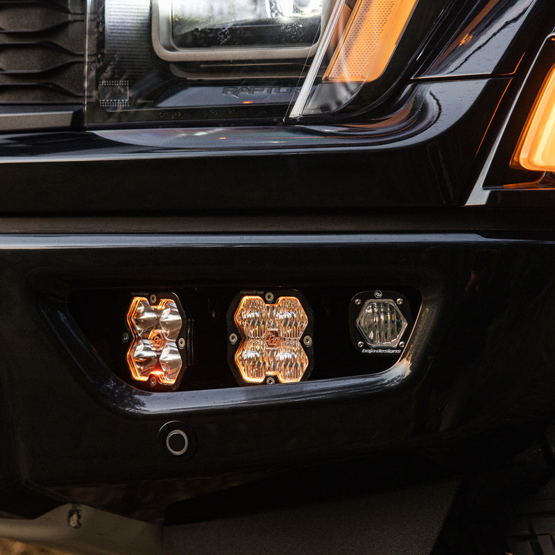 Baja Designs Squadron 2.0 Pro/S1 Fog Pocket Light Kit on a Ford F-150 Raptor: two square pods with amber backlights and a small S1 pod installed in the fog pocket of a black Ford Raptor