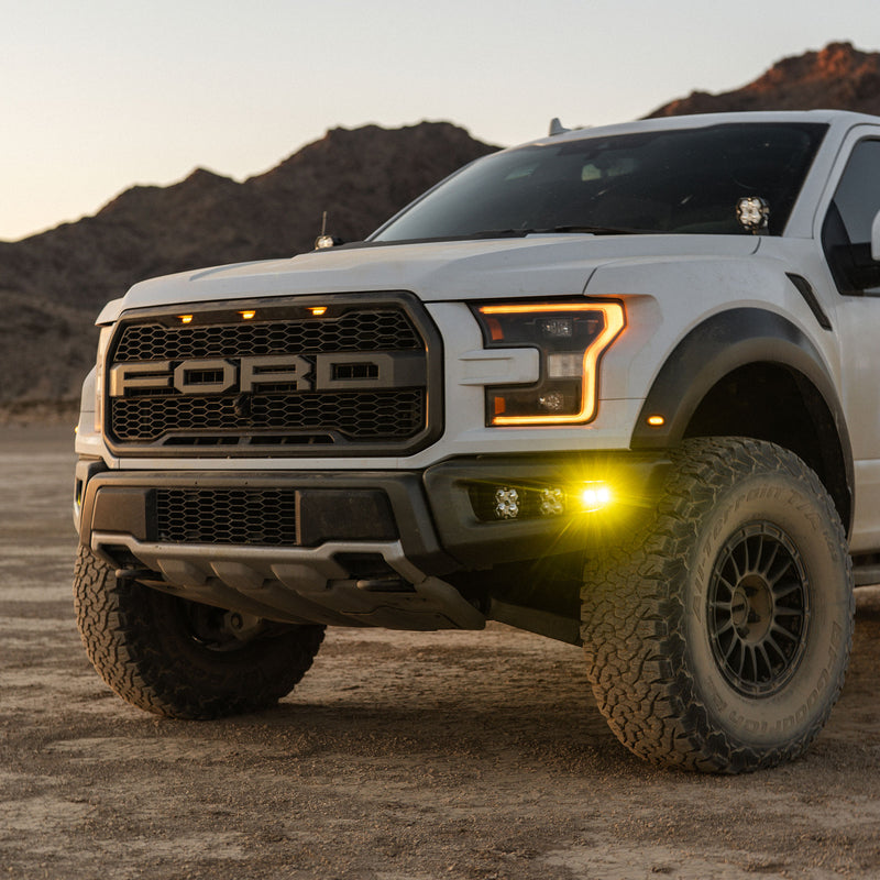 Baja Designs Squadron 2.0 Pro/S2 SAE Pro Fog Pocket Light Kit installed on a Ford F-150 Raptor: white Ford Raptor pickup in the desert with amber fog pocket lights lit in the front bumper