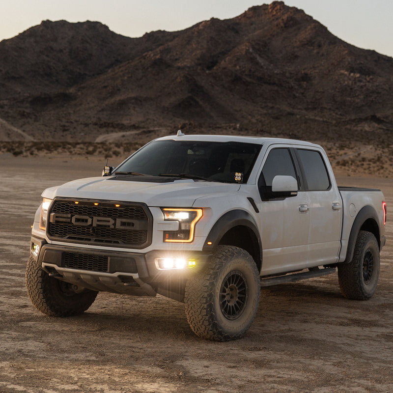 Baja Designs Squadron 2.0 Pro/S2 SAE Pro Fog Pocket Light Kit installed on a Ford F-150 Raptor: white Ford Raptor pickup on a desert flat at dusk with white fog pocket lights lit