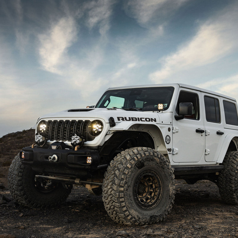 Baja Designs Squadron 2.0 Sport A-Pillar Light Kit installed on a Jeep Wrangler: A lifted white four-door Jeep Wrangler Rubicon on rocky ground with pods mounted at the A-pillars and lights on the front bumper