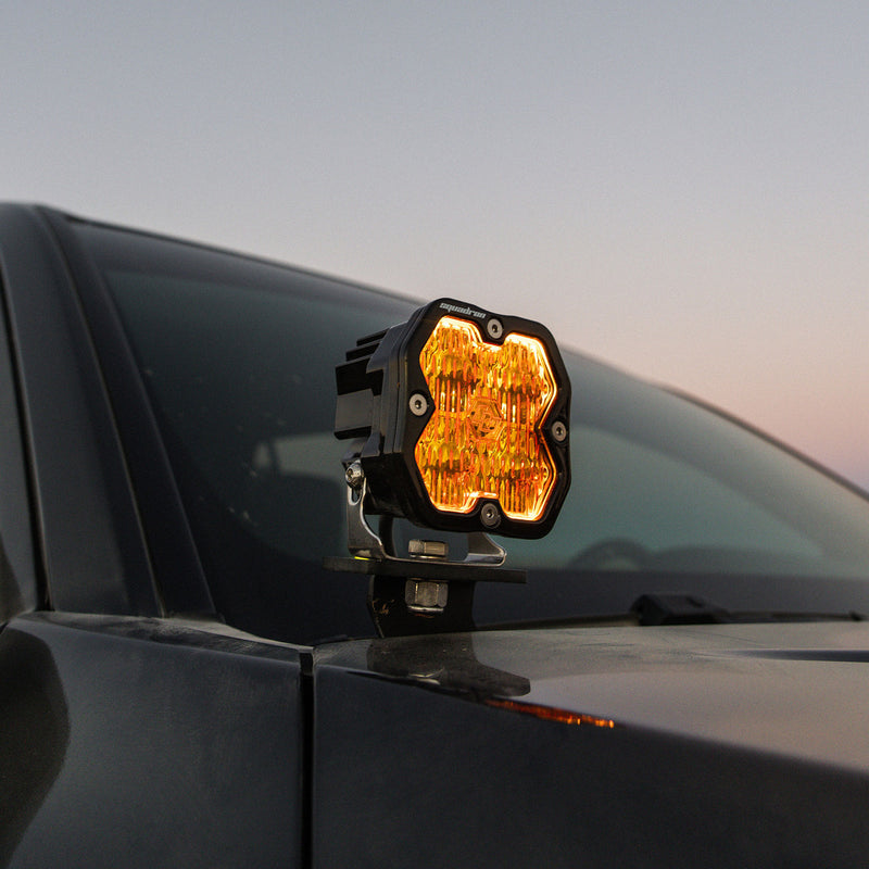 Baja Designs Squadron 2.0 Sport A-Pillar Light Kit on a vehicle: A Squadron pod glowing amber on a black A-pillar mount beside the windshield of a dark vehicle at dusk