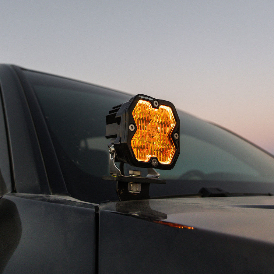 A-Pillar Light Kits by Baja Designs: A Squadron pod glowing amber on a black A-pillar mount beside the windshield of a dark vehicle at dusk