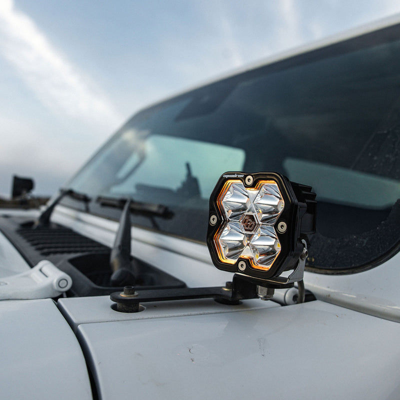 Baja Designs Squadron 2.0 Sport A-Pillar Light Kit on a Jeep Wrangler: A Squadron pod with a glowing amber backlight on an A-pillar mount at the windshield of a white Jeep