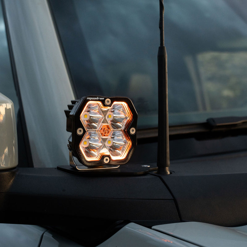 Baja Designs Squadron 2.0 Sport A-Pillar Light Kit on a vehicle: A Squadron pod with a glowing amber backlight on a cowl mount beside an antenna at the windshield of a gray vehicle