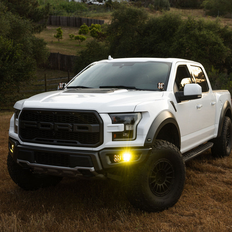 Baja Designs Squadron 2.0 Sport A-Pillar Light Kit installed on a Ford F-150 Raptor: A white Ford Raptor pickup at dusk with pods on both A-pillars and amber fog lights glowing in the bumper