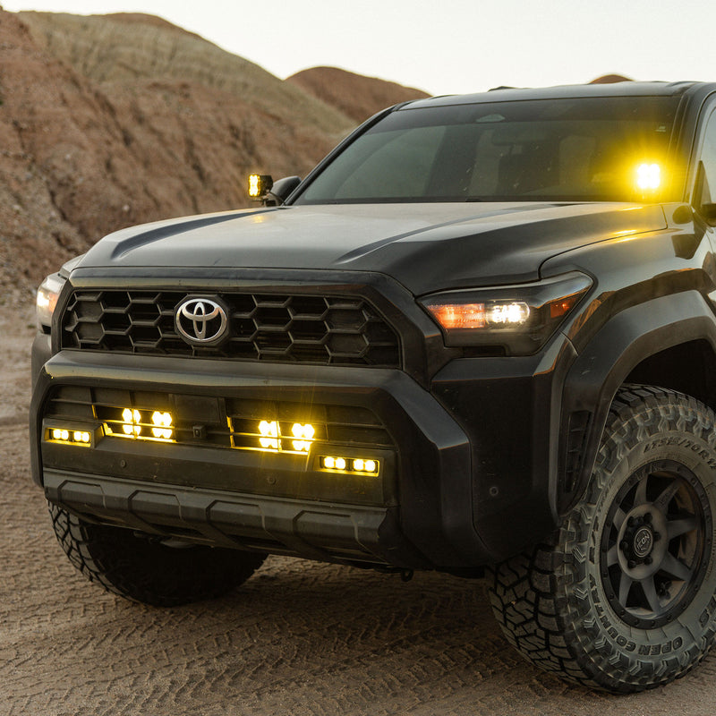 Baja Designs Squadron 2.0 Sport Behind-the-Grille Light Kit installed on a Toyota: a dark Toyota on a dirt trail with amber pods glowing behind the lower grille and on the A-pillars