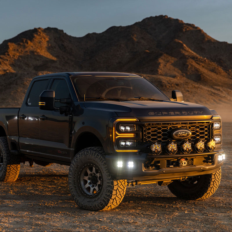 Baja Designs Squadron 2.0 Sport Dual Fog Pocket Light Kit installed on a Ford F-250/F-350 Super Duty: black Ford Super Duty pickup in the desert at dusk with lit square fog pocket lights and a row of pods across the grille