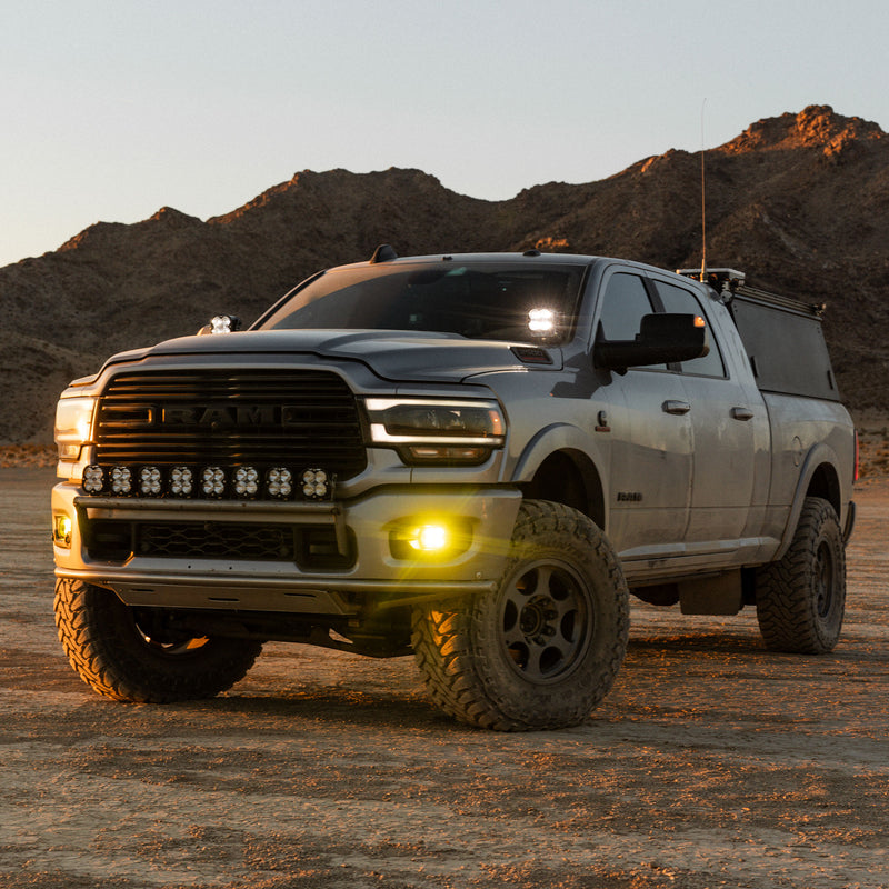 Baja Designs Squadron 2.0 Sport Fog Pocket Light Kit installed on a Ram 2500: gray Ram 2500 pickup in the desert at dusk with a lit amber fog pocket light, a row of bumper pods and pillar lights