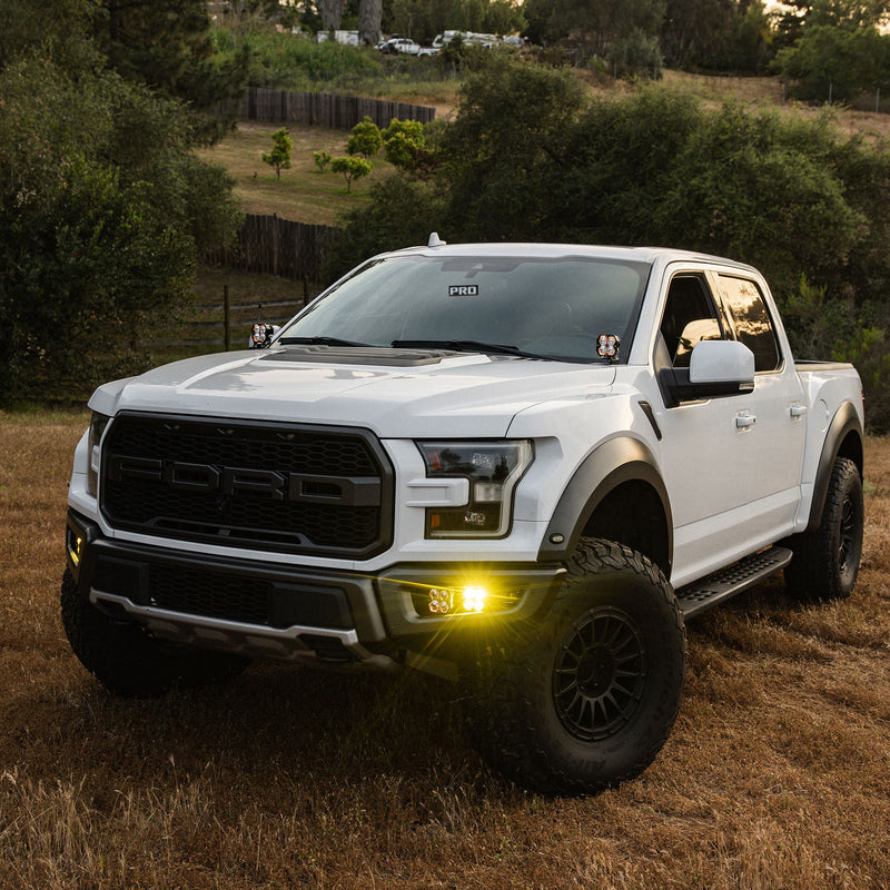 Baja Designs Squadron 2.0 Sport/S2 Sport Fog Pocket Light Kit installed on a Ford F-150 Raptor: white Ford Raptor pickup parked on dry grass with amber fog pocket lights lit