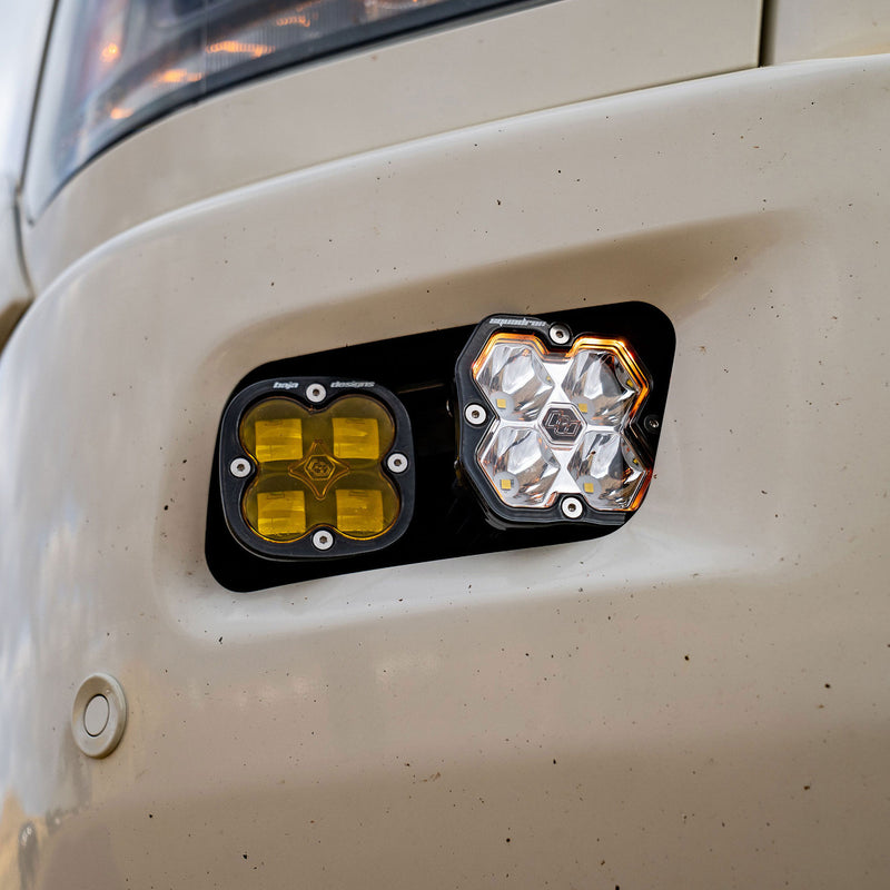 Baja Designs Squadron 2.0 Sport/Squadron SAE Dual Fog Pocket Light Kit on a vehicle: close view of an amber and a clear square Squadron pod mounted in a black dual fog pocket bezel on a white bumper