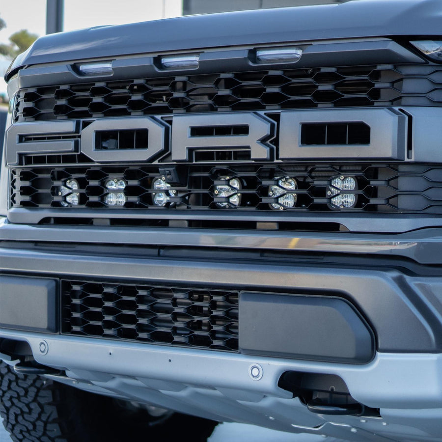 Baja Designs Squadron Pro Behind Grille Light Kit on a Ford F-150 Raptor: gray Ford Raptor grille with six square LED pods lit behind the lower grille below the FORD lettering
