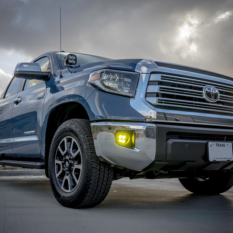 Baja Designs Squadron-R Sport Fog Pocket Light Kit installed on a Toyota Tundra: A blue Toyota Tundra under a stormy sky with an amber fog light glowing in the front bumper pocket