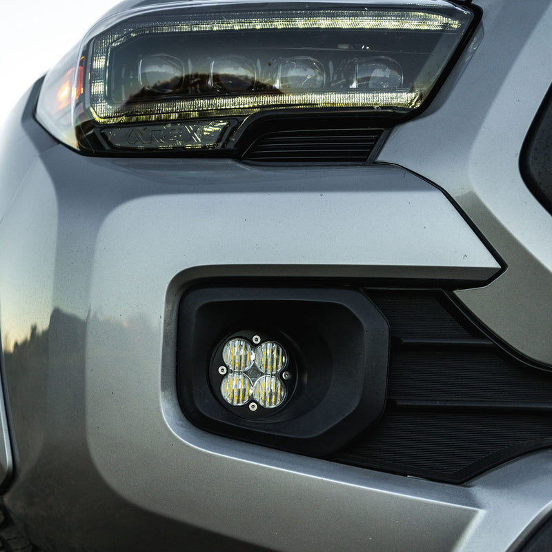 Baja Designs Squadron-R Sport Fog Pocket Light Kit on a vehicle: close view of a clear round Squadron fog light fitted in the fog pocket of a silver vehicle below the headlight