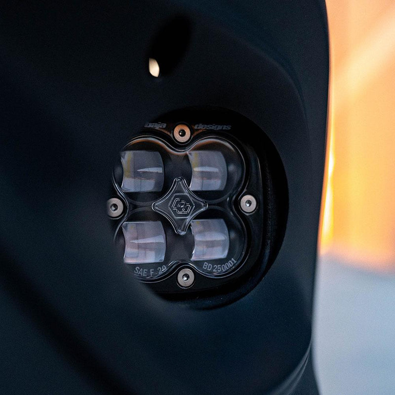 Baja Designs Squadron SAE Fog Pocket Light Kit on a vehicle: black Squadron SAE pod with a clear lens set into a round fog pocket on a dark vehicle bumper