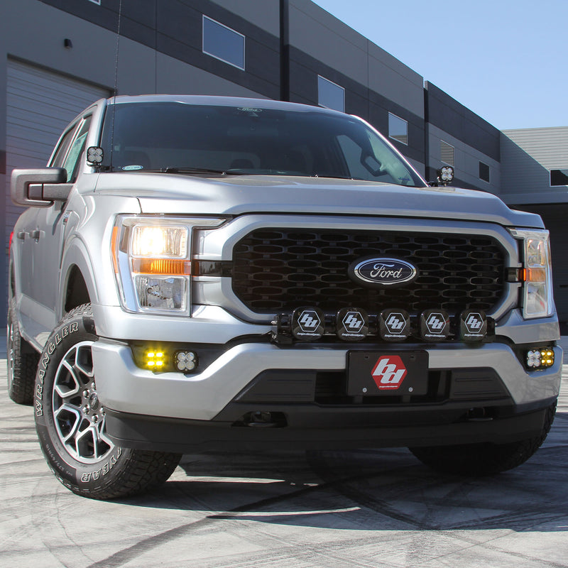 Baja Designs Squadron SAE/Pro Fog Pocket Light Kit installed on a Ford F-150: A silver Ford F-150 parked by a building with A-pillar pods, a row of grille pods and amber fog lights