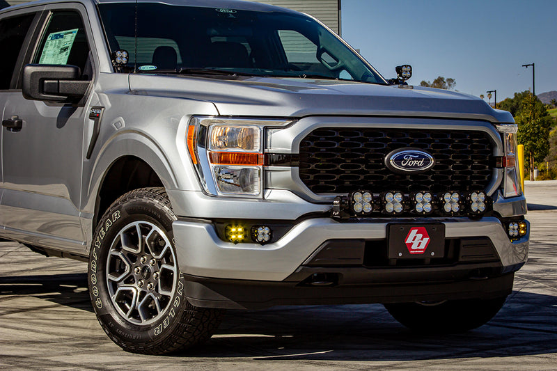 Baja Designs Squadron SAE/Sport Fog Pocket Light Kit installed on a Ford F-150: silver Ford F-150 front three-quarter view with lit amber fog pocket pods, a pod bar in the lower grille and a Baja Designs logo plate; amber may not match the clear